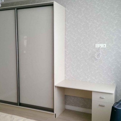 шкаф купе в спальню с письменным столом Шкаф-купе в интерьере спальни. Справа от шкафа находится письменный стол. Вся мебель в светлых тонах.